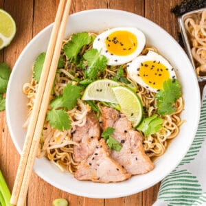 Square image of Instant Pot Ramen topped with sliced pork, soft-boiled eggs, bean sprouts, cilantro, lime wedges, black sesame seeds, and chopsticks