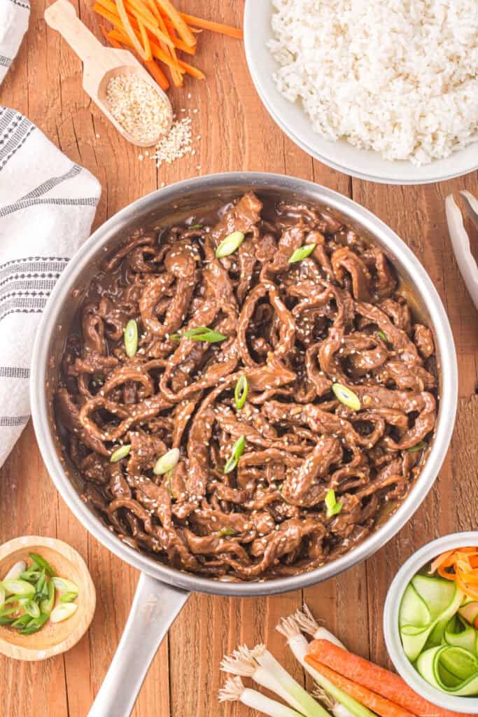 Korean Beef Bulgogi in a skillet topped with sesame seeds and green onions served with white rice and vegetables