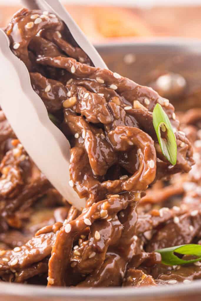 Close-up of Korean Beef Bulgogi being lifted with tongs showing thick glossy bulgogi sauce and sesame seeds