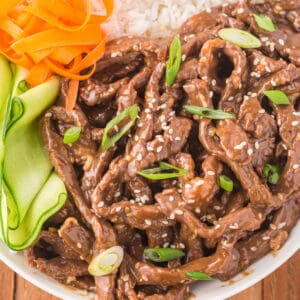 Square image of Korean Beef Bulgogi served with white rice, cucumber ribbons, carrots, sesame seeds, and green onions