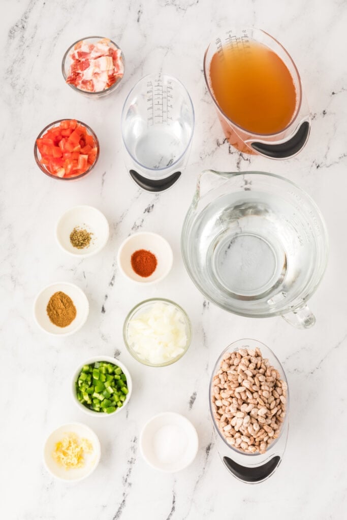 Ingredients for Ranchero Beans including pinto beans, bacon, broth, tomatoes, onion, jalapeño, and spices on a marble surface