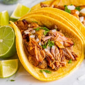 Square image of Slow Cooker Carnitas taco with lime wedges and cilantro