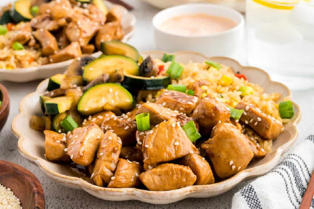 Plate of homemade Hibachi Chicken served with fried rice, zucchini, mushrooms, yum yum sauce, and sesame seeds for a takeout-style meal at home.