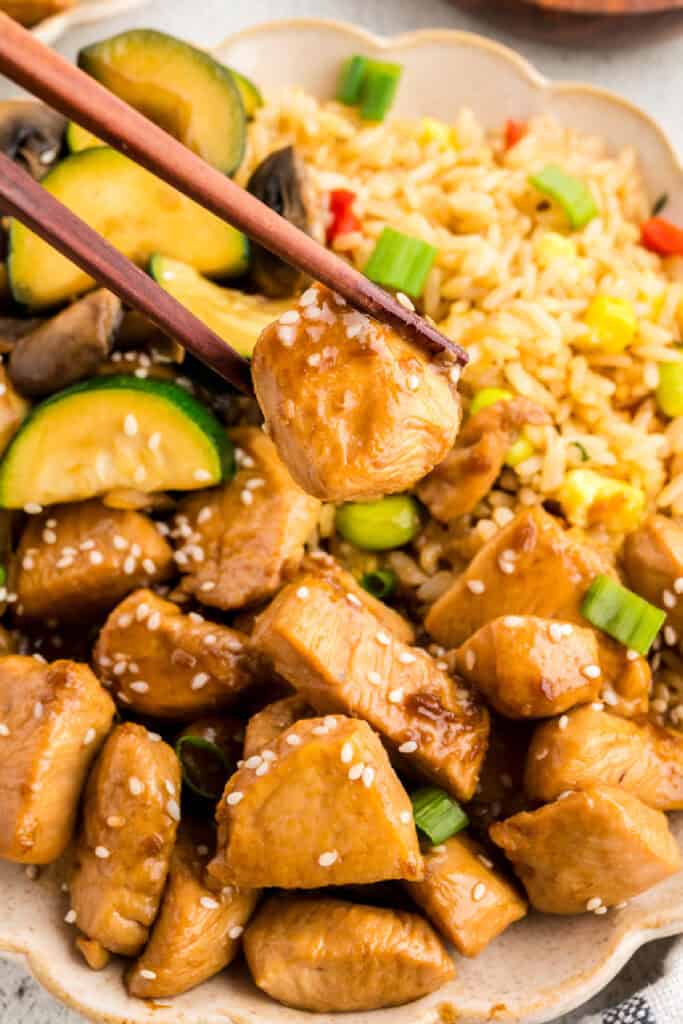 Chopsticks holding a piece of Hibachi Chicken over fried rice and hibachi vegetables with sesame seeds and green onions.