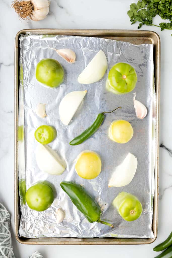 raw tomatillos, onion wedges, jalapeño, and garlic arranged on a foil-lined baking sheet before roasting for Tomatillo Salsa
