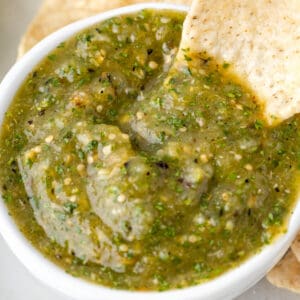 tortilla chip dipping into a bowl of Tomatillo Salsa with visible herbs, seeds, and slightly chunky texture