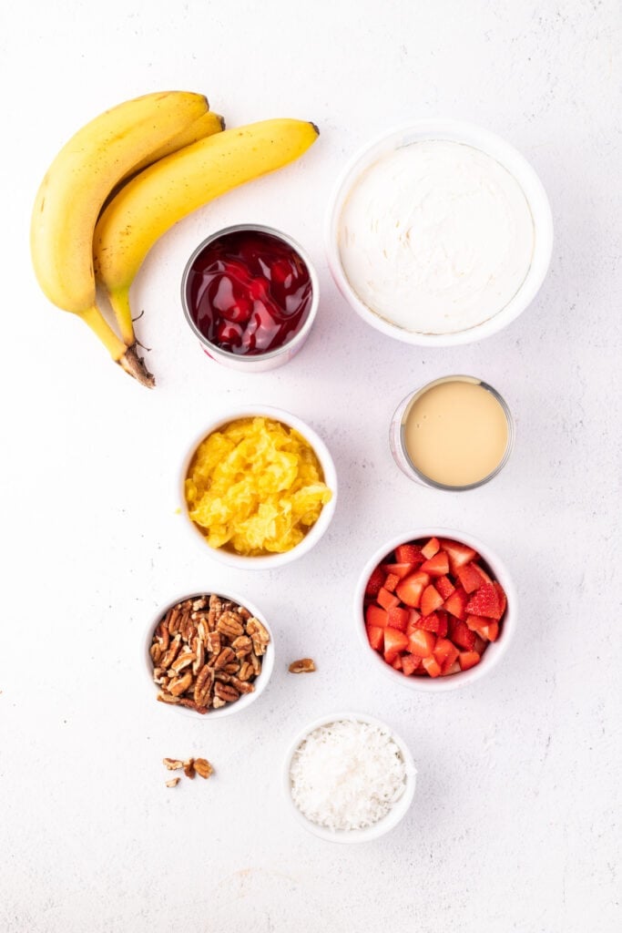 Ingredients for Banana Split Salad arranged on a white surface, including bananas, cherry pie filling, whipped topping, sweetened condensed milk, crushed pineapple, strawberries, pecans, and coconut.