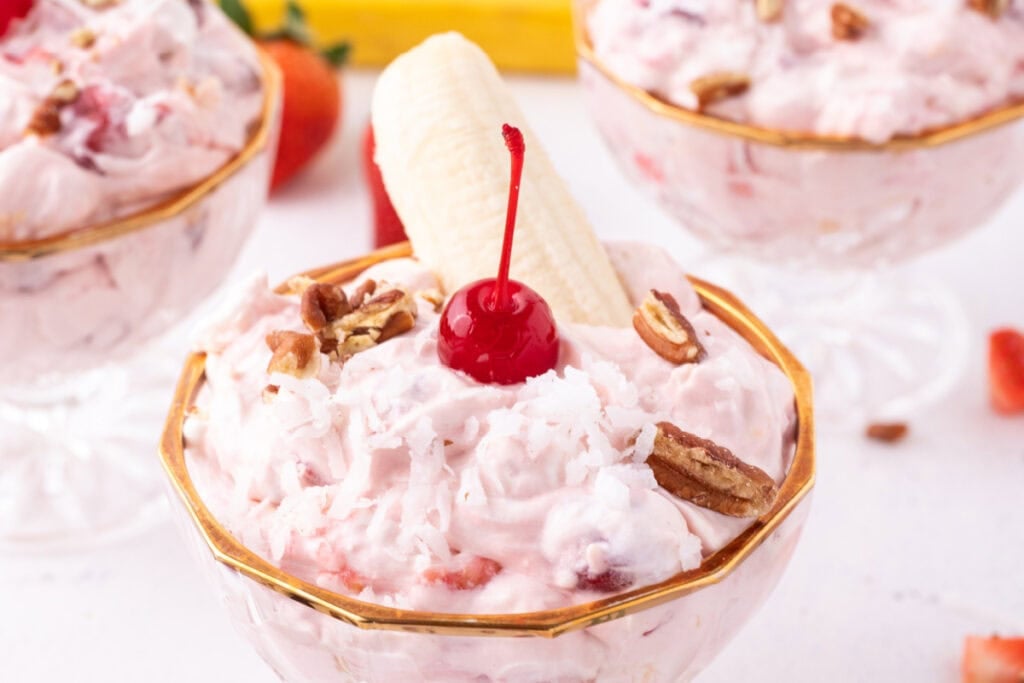 Individual serving of Banana Split Salad topped with coconut, pecans, a maraschino cherry, and a banana half in a dessert dish.