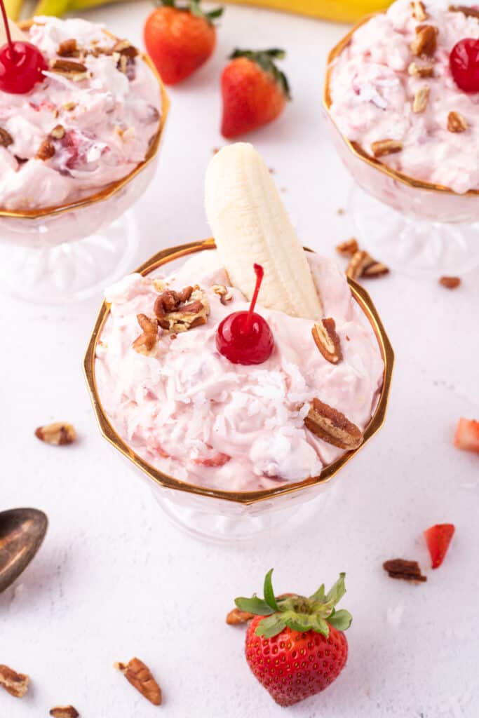 Three dessert dishes filled with Banana Split Salad and garnished with bananas, pecans, coconut, and maraschino cherries.