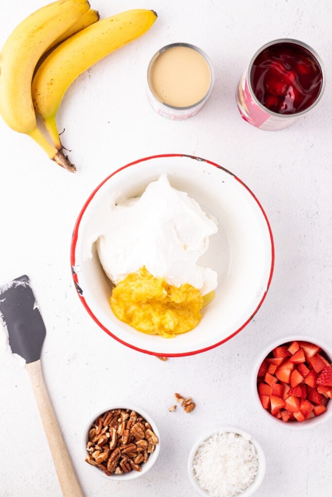 Whipped topping and crushed pineapple in a mixing bowl with bananas, sweetened condensed milk, cherry pie filling, strawberries, pecans, and coconut nearby for Banana Split Salad.