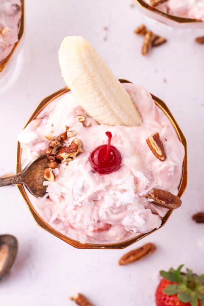Overhead close-up of Banana Split Salad topped with coconut, pecans, a maraschino cherry, and a banana half in a dessert dish.