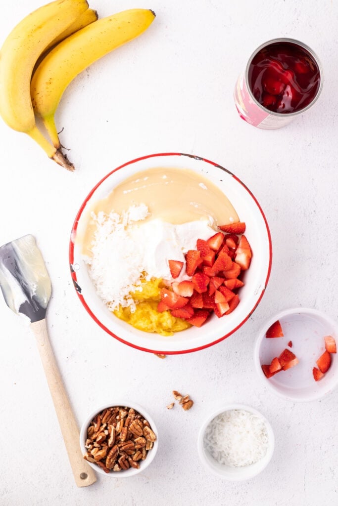 Ingredients for Banana Split Salad in a mixing bowl, including whipped topping, sweetened condensed milk, crushed pineapple, coconut, and chopped strawberries.