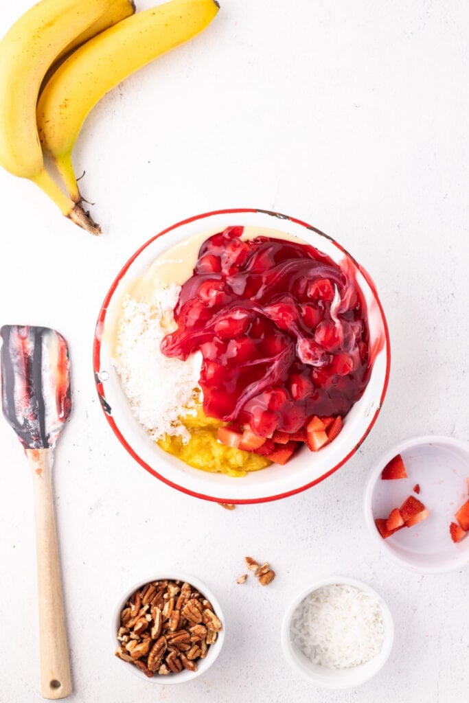 Cherry pie filling added to a mixing bowl with whipped topping, sweetened condensed milk, pineapple, strawberries, and coconut for Banana Split Salad.