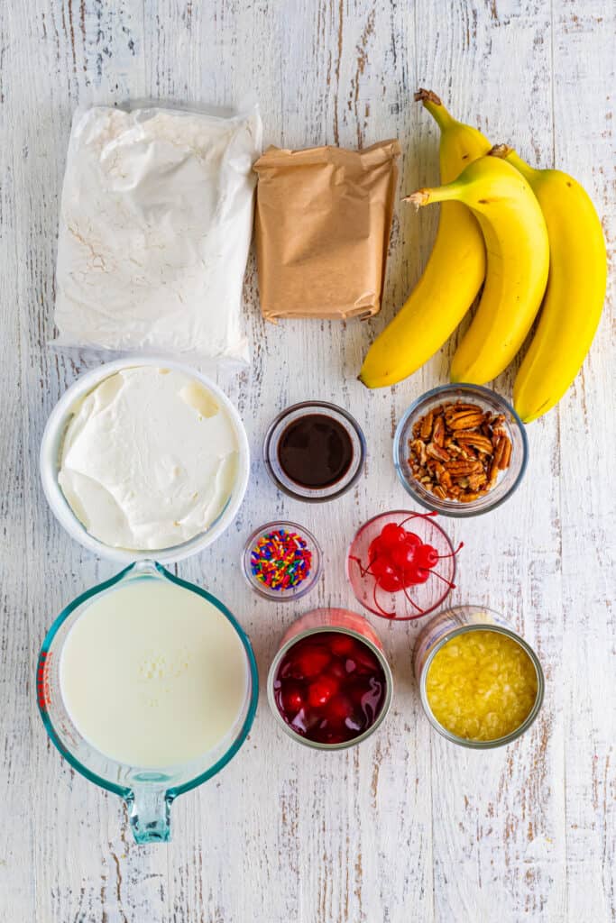 Ingredients for Banana Split Trifle arranged on a white wood surface, including yellow cake mix, vanilla pudding mix, whole milk, bananas, whipped topping, crushed pineapple, strawberry pie filling, chocolate sauce, pecans, rainbow sprinkles, and maraschino cherries.