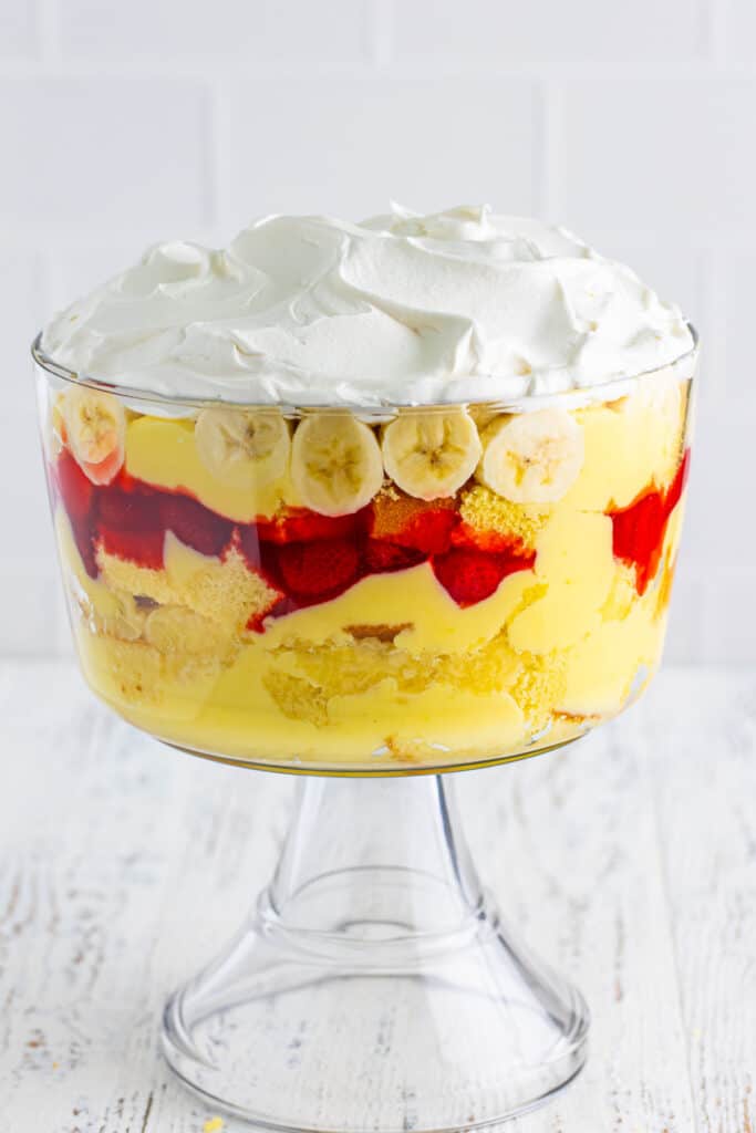 Whipped topping spread over sliced bananas in a clear trifle bowl for Banana Split Trifle.