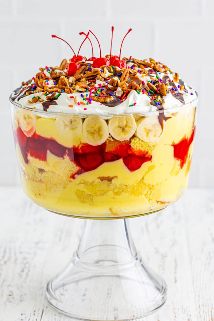 Finished Banana Split Trifle topped with whipped topping, chocolate sauce, chopped pecans, rainbow sprinkles, and maraschino cherries in a clear trifle bowl.