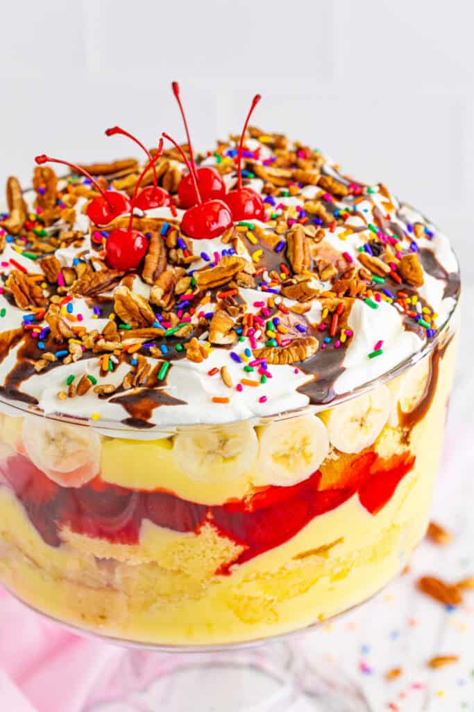Close-up of finished Banana Split Trifle showing layers of cake, pudding, pineapple, strawberry filling, bananas, whipped topping, chocolate sauce, pecans, sprinkles, and cherries.