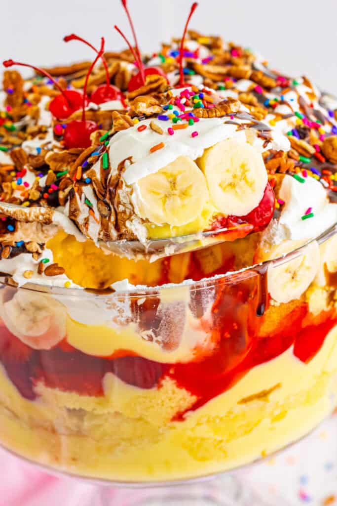 Spoon lifting a serving of Banana Split Trifle from the trifle bowl, showing layers of cake, pudding, bananas, strawberry filling, whipped topping, chocolate sauce, pecans, sprinkles, and cherries.