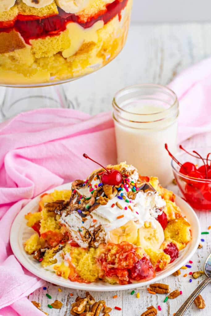 Serving of Banana Split Trifle on a plate topped with whipped topping, chocolate sauce, pecans, rainbow sprinkles, and a maraschino cherry, with the trifle bowl in the background.