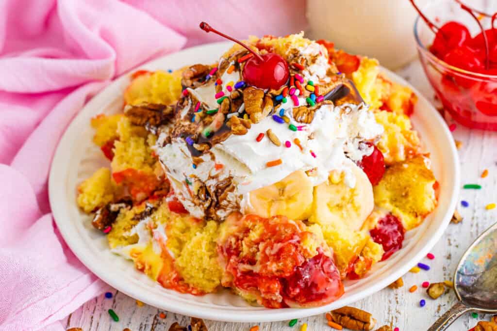 Close-up of a plated serving of Banana Split Trifle topped with whipped topping, chocolate sauce, pecans, rainbow sprinkles, and a maraschino cherry.