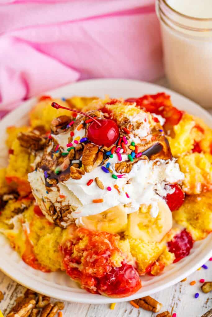 Close-up of Banana Split Trifle on a plate with layers of cake, pudding, strawberry filling, bananas, whipped topping, chocolate sauce, pecans, rainbow sprinkles, and a maraschino cherry.