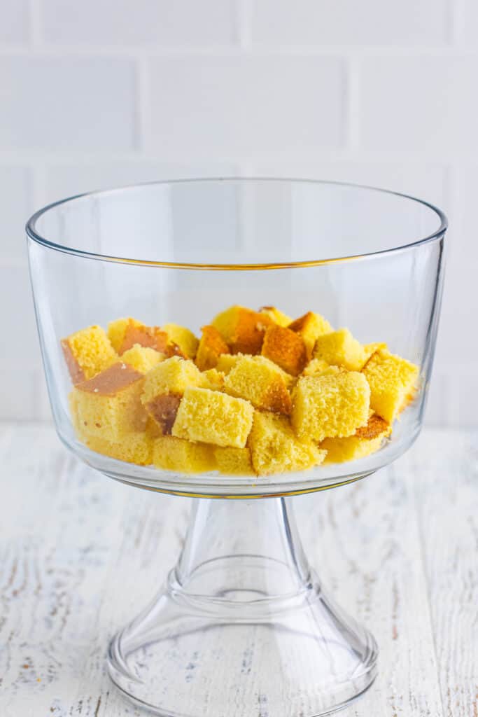 Cubed yellow cake layered in the bottom of a clear trifle bowl for Banana Split Trifle.