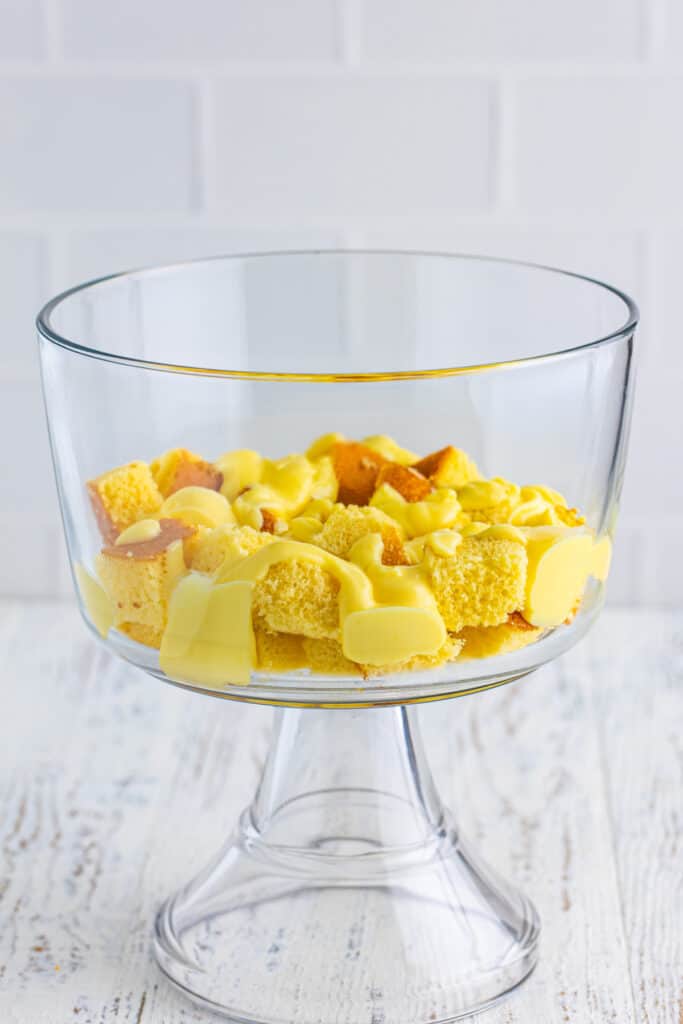 Cubed yellow cake topped with vanilla pudding in a clear trifle bowl for Banana Split Trifle.