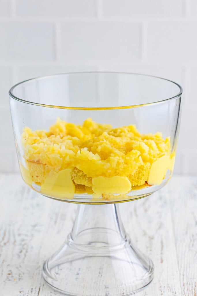 Layer of crushed pineapple spread over cake and pudding in a clear trifle bowl for Banana Split Trifle.