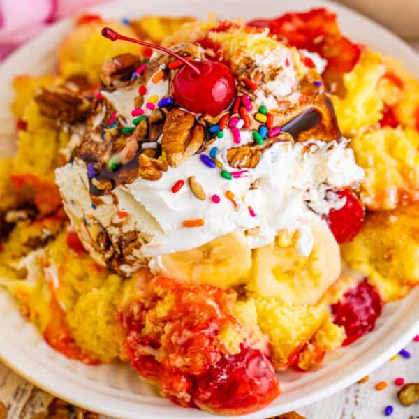 Square image of Banana Split Trifle topped with whipped topping, chocolate sauce, pecans, rainbow sprinkles, and maraschino cherries, showing the colorful dessert layers.
