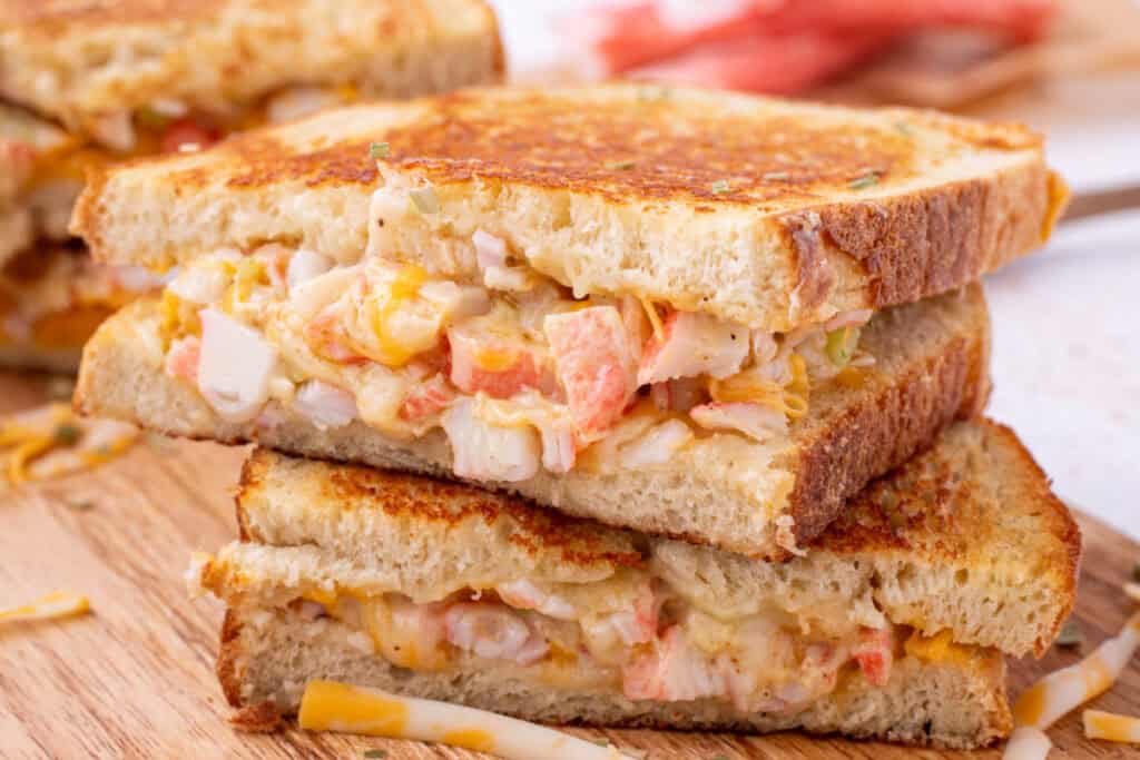 Four Cheesy Crab Melt sandwich halves stacked on a wooden cutting board showing the crab filling