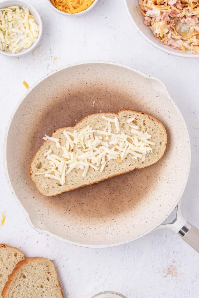 Shredded Gruyere cheese sprinkled onto a slice of sourdough bread in a skillet