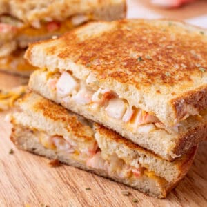 Square image of stacked Cheesy Crab Melt sandwich halves on a wooden cutting board