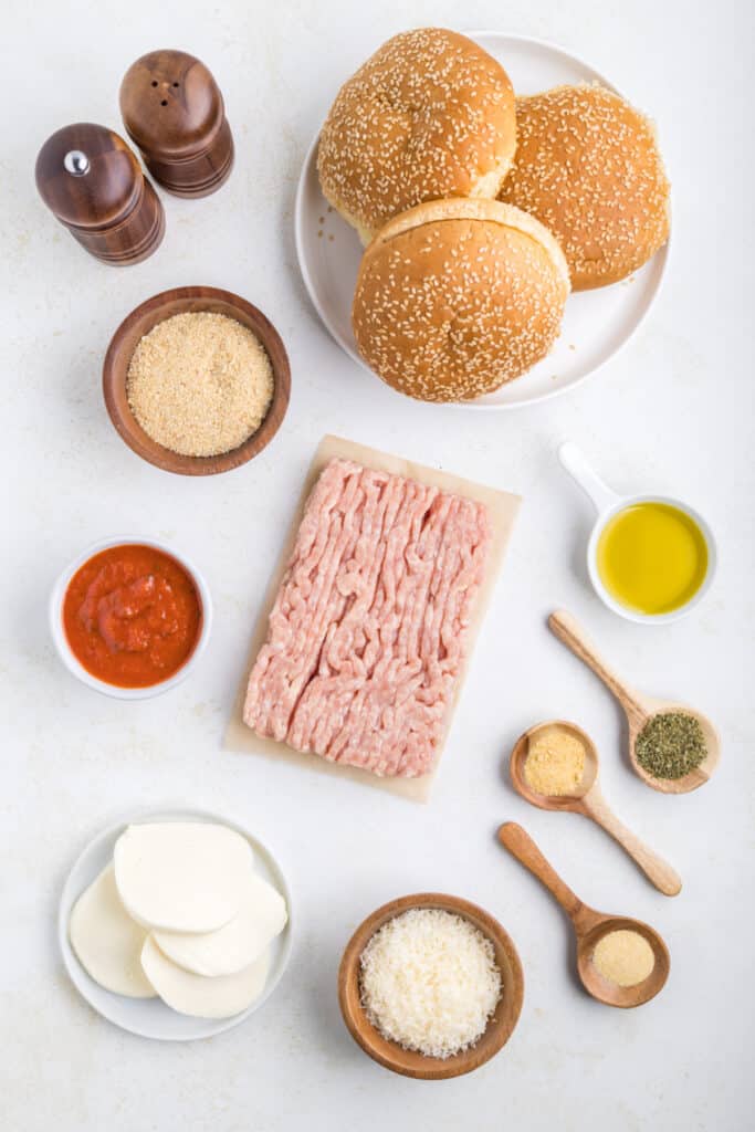 Ingredients for Chicken Parm Burgers arranged on a countertop including ground chicken, marinara sauce, breadcrumbs, Parmesan cheese, mozzarella, seasonings, olive oil, and burger buns