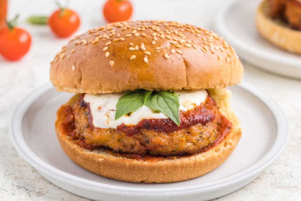Close-up of a Chicken Parm Burger with marinara sauce, melted mozzarella, and basil on a sesame seed bun