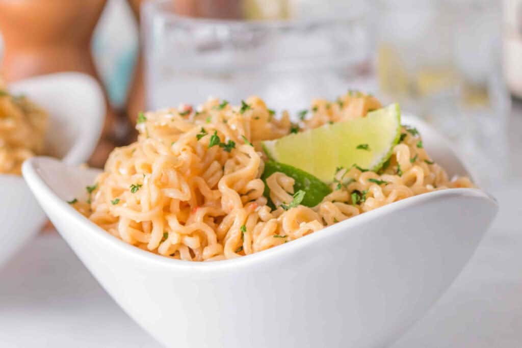 Creamy Peanut Butter Ramen Noodles served in a white bowl with sesame seeds, parsley, lime wedges, and chopsticks.