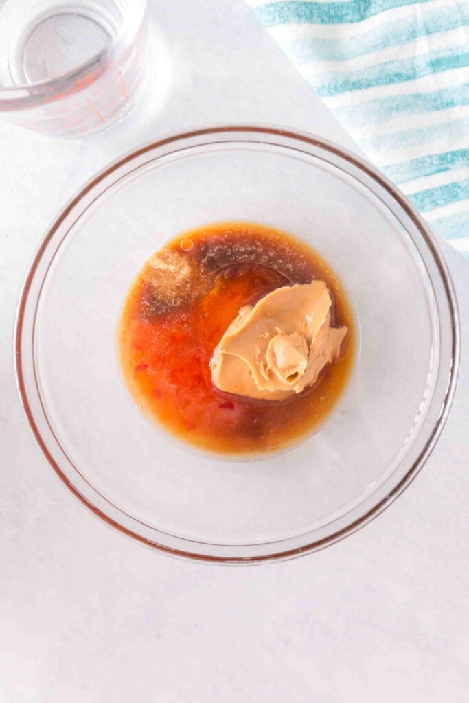 Peanut butter, sriracha, Worcestershire sauce, brown sugar, garlic powder, lime juice, olive oil, and water combined in a mixing bowl before whisking.