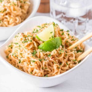 Creamy Peanut Butter Ramen Noodles with sesame seeds, parsley, lime wedges, and chopsticks in a white serving bowl.