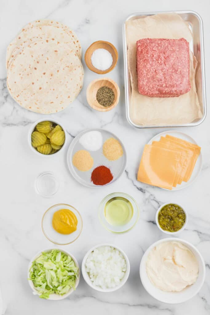 Ingredients for Smash Burger Tacos arranged on a marble surface, including flour tortillas, ground beef, American cheese, lettuce, onion, pickles, mayonnaise, relish, mustard, oil, and seasonings.