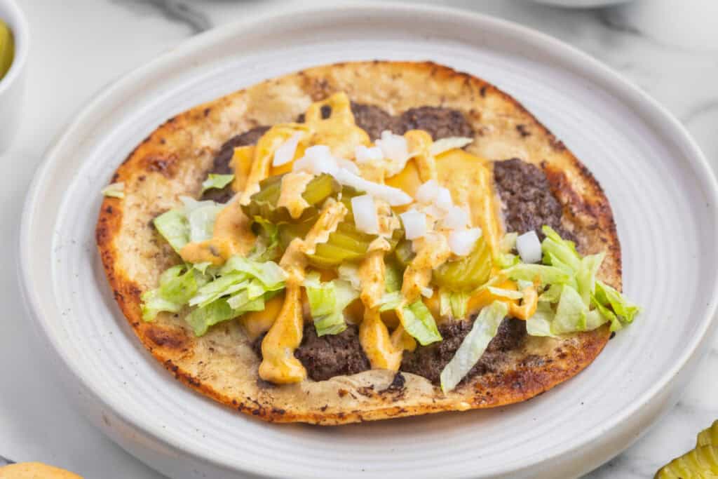 Smash Burger Taco on a plate topped with melted American cheese, burger sauce, shredded lettuce, diced onion, and pickle slices.