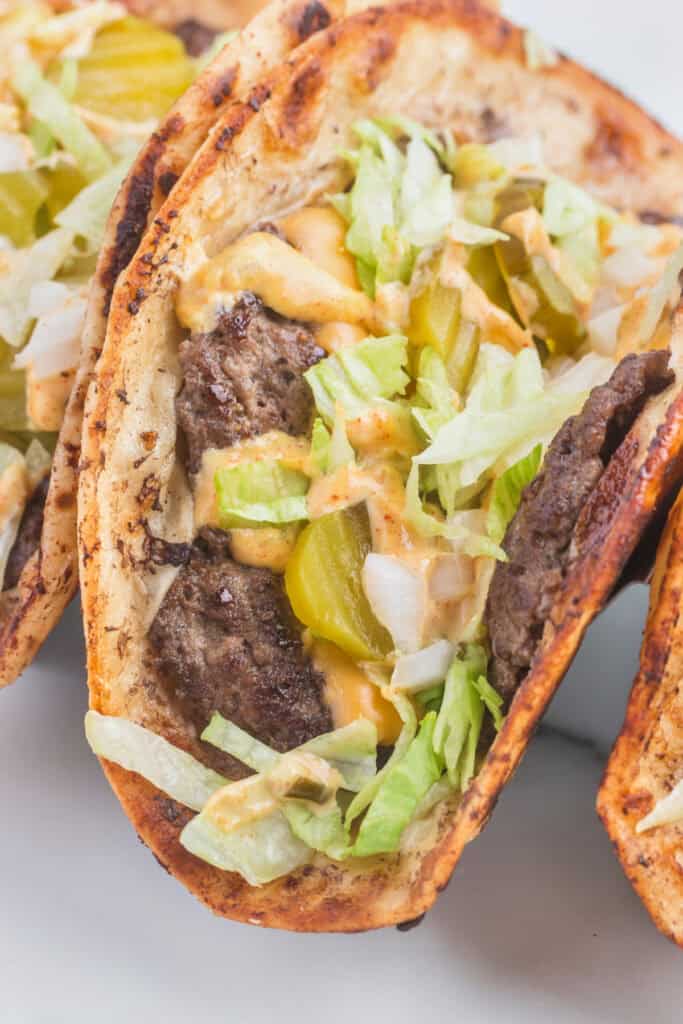 Close-up of a folded Smash Burger Taco filled with seasoned beef, melted cheese, burger sauce, shredded lettuce, diced onion, and pickles.