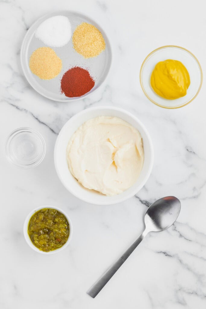 Bowl of mayonnaise with pickle relish, mustard, seasonings, vinegar, and a spoon arranged nearby for making burger sauce.