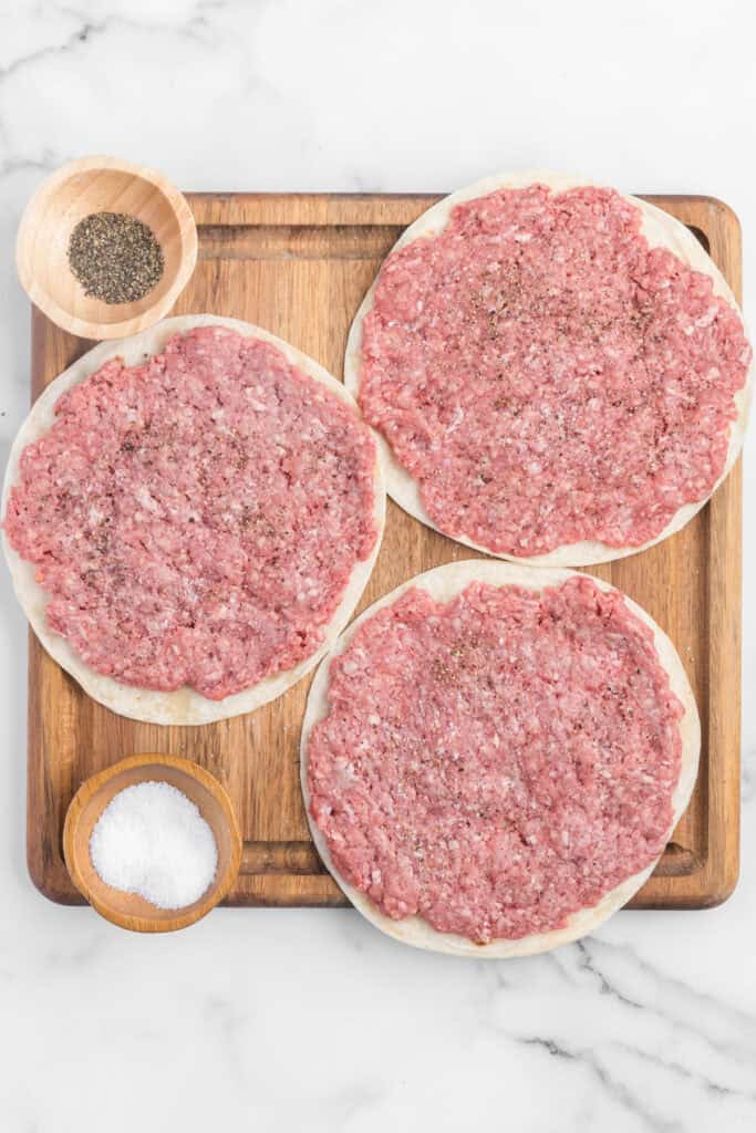 Flour tortillas topped with a thin layer of raw ground beef and seasoned with salt and pepper on a wooden cutting board.