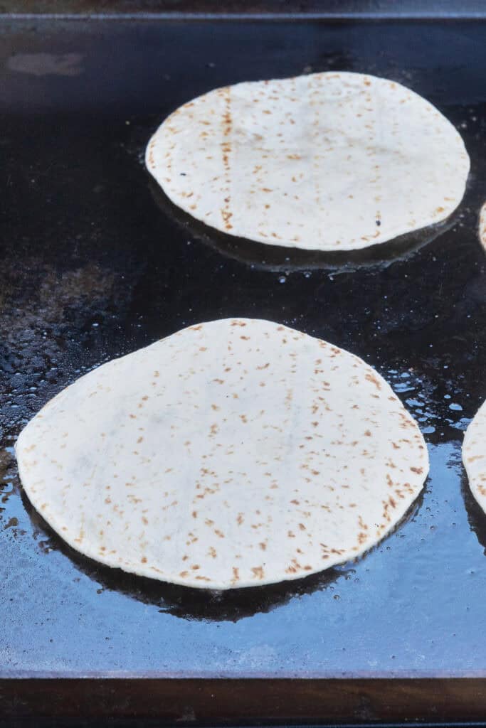 Flour tortillas cooking on a flat-top griddle before the ground beef side is browned.