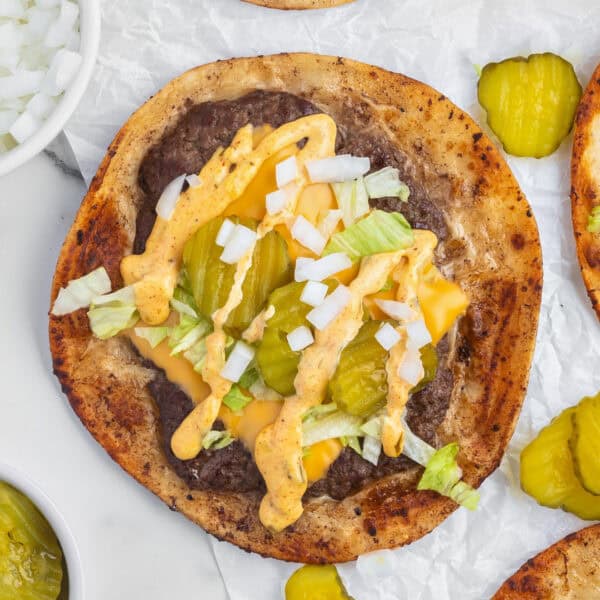 Overhead view of a Smash Burger Taco topped with melted cheese, burger sauce, shredded lettuce, diced onion, and pickle slices on parchment paper.