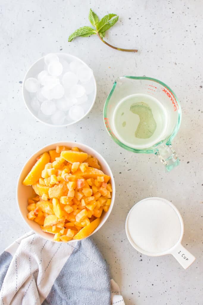 Ingredients for a Frozen Peach Bellini including frozen peaches, granulated sugar, ice, Prosecco, lemon juice, and fresh mint.