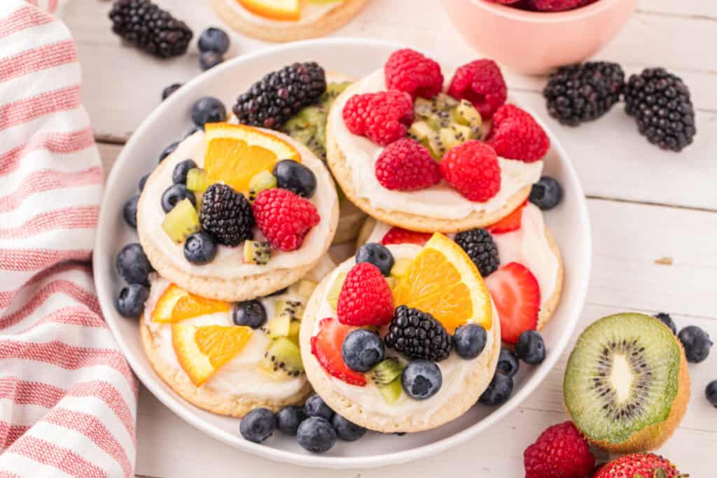 Platter of homemade Fruit Pizza Cookies topped with cream cheese frosting, fresh berries, kiwi, and orange slices.