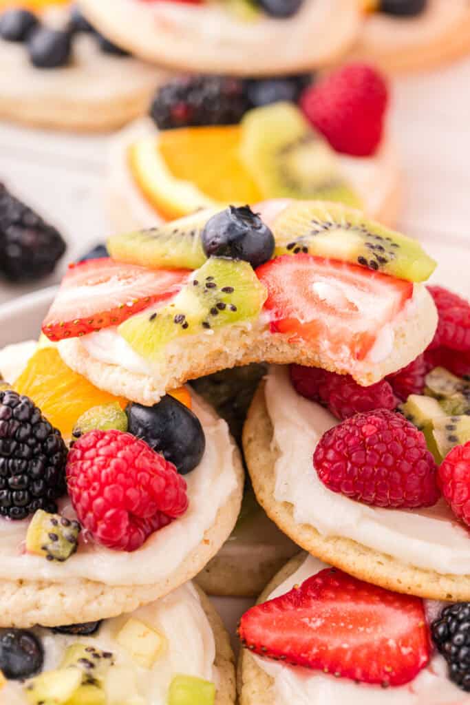 Bitten Fruit Pizza Cookie topped with cream cheese frosting, strawberries, kiwi, and blueberries, showing the soft sugar cookie center.
