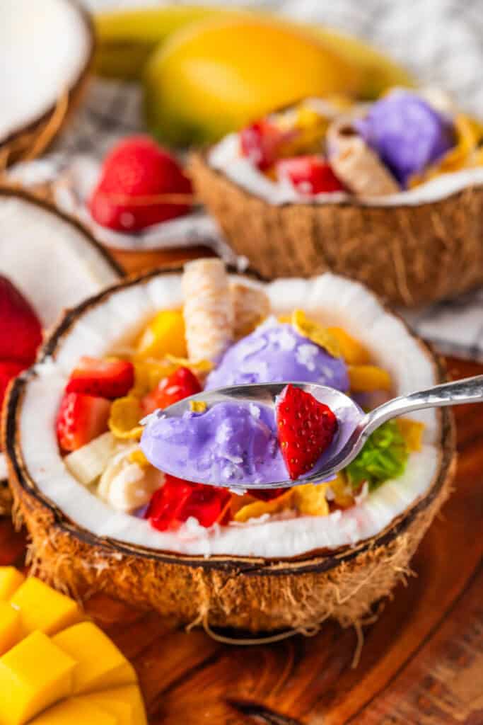 Spoonful of Halo Halo with creamy ube ice cream and fresh strawberry above a coconut bowl filled with tropical toppings.