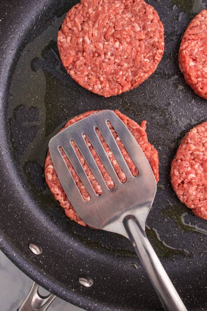 Smashing burger patties thin with a spatula in a hot skillet.