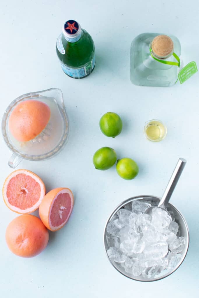 Ingredients for a fresh Paloma Cocktail arranged on a countertop with grapefruit, limes, tequila, sparkling water, agave syrup, and ice.